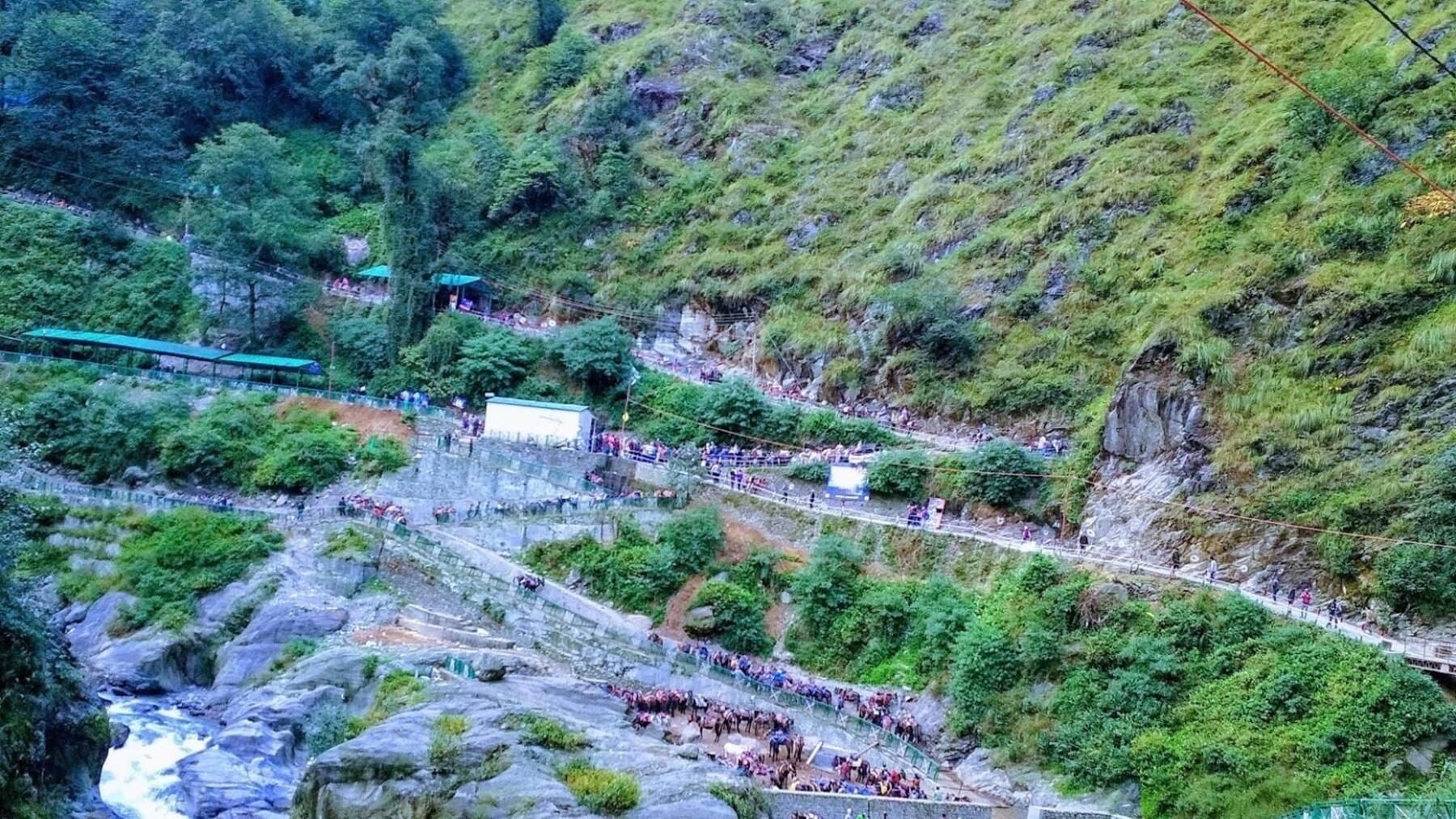 A paved trail with railings climbing a green valley side, with a rocky stream below and pilgrims walking along it
